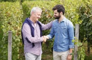 Older man shaking a younger mans hand with grape vines in the background.